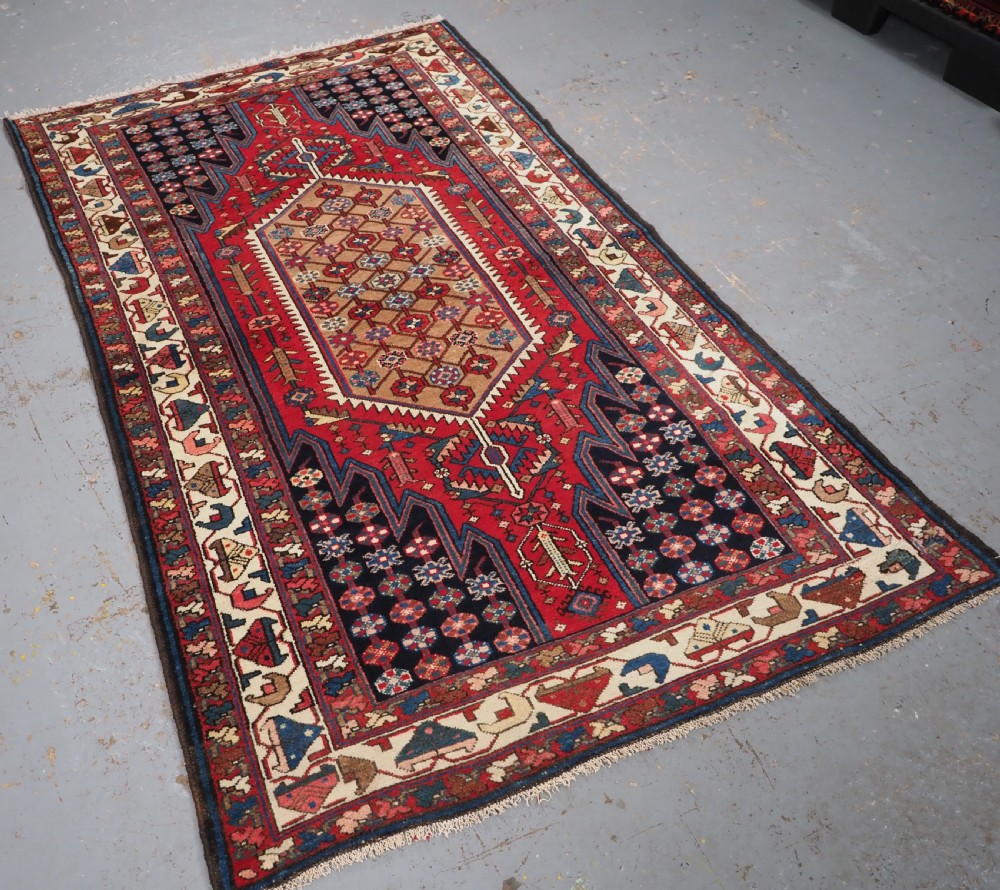 An antique hand-knotted Persian Mazlaghan wool rug featuring a central camel-coloured medallion on a red stepped field with dark blue rosette borders. View II