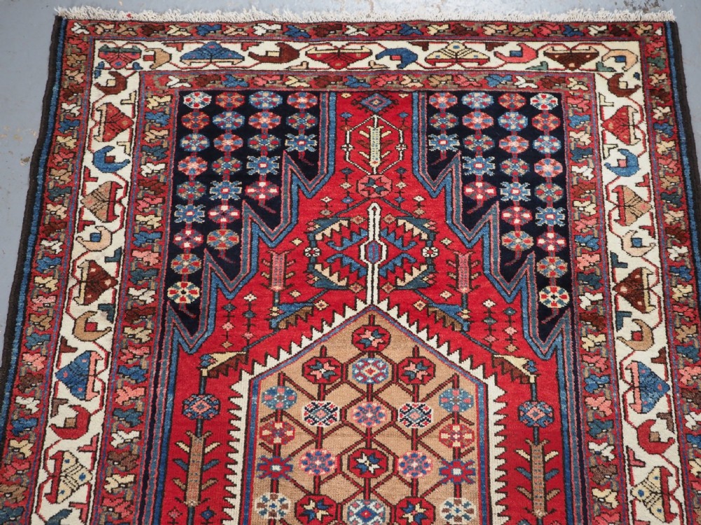 An antique hand-knotted Persian Mazlaghan wool rug featuring a central camel-coloured medallion on a red stepped field with dark blue rosette borders. View III