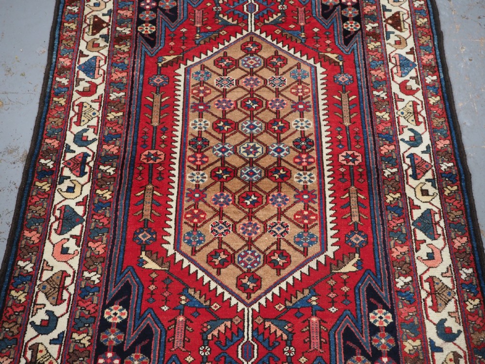 An antique hand-knotted Persian Mazlaghan wool rug featuring a central camel-coloured medallion on a red stepped field with dark blue rosette borders. View IV
