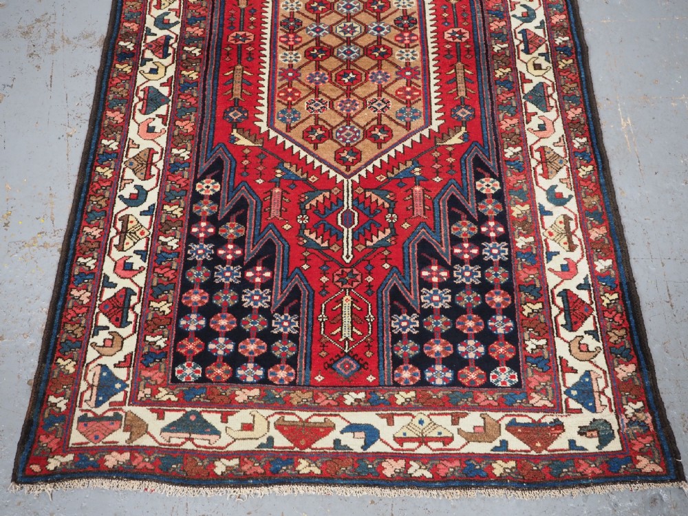 An antique hand-knotted Persian Mazlaghan wool rug featuring a central camel-coloured medallion on a red stepped field with dark blue rosette borders. View V