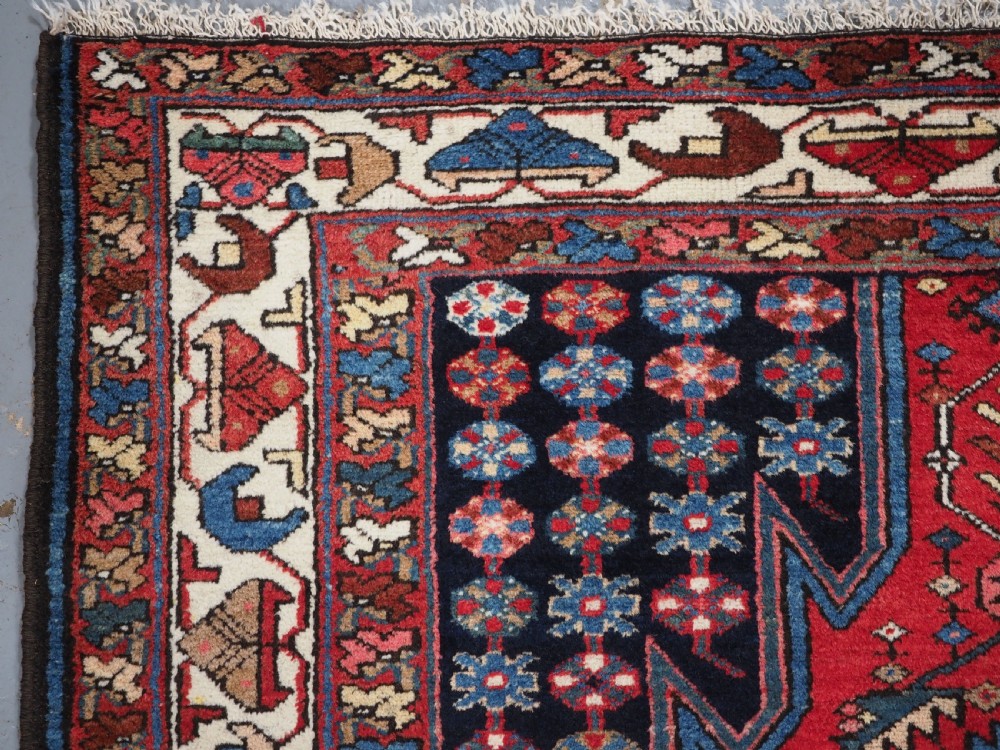 An antique hand-knotted Persian Mazlaghan wool rug featuring a central camel-coloured medallion on a red stepped field with dark blue rosette borders. View VI