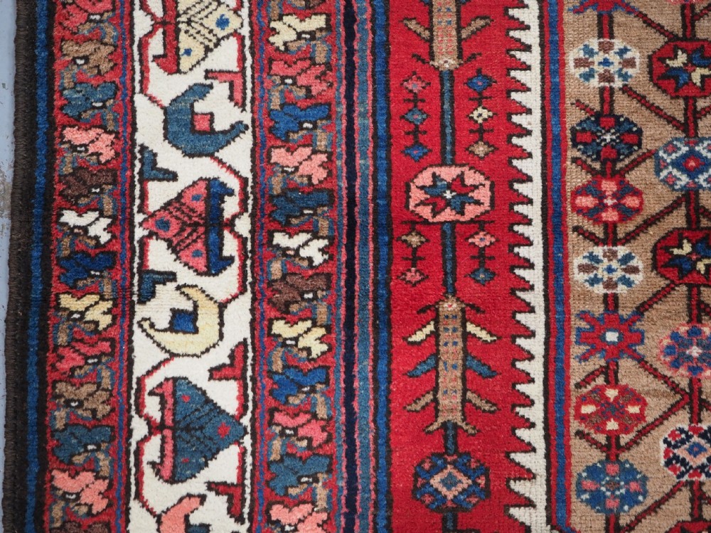 An antique hand-knotted Persian Mazlaghan wool rug featuring a central camel-coloured medallion on a red stepped field with dark blue rosette borders. View VII