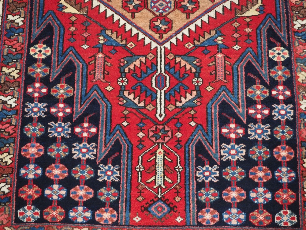 An antique hand-knotted Persian Mazlaghan wool rug featuring a central camel-coloured medallion on a red stepped field with dark blue rosette borders. View IX