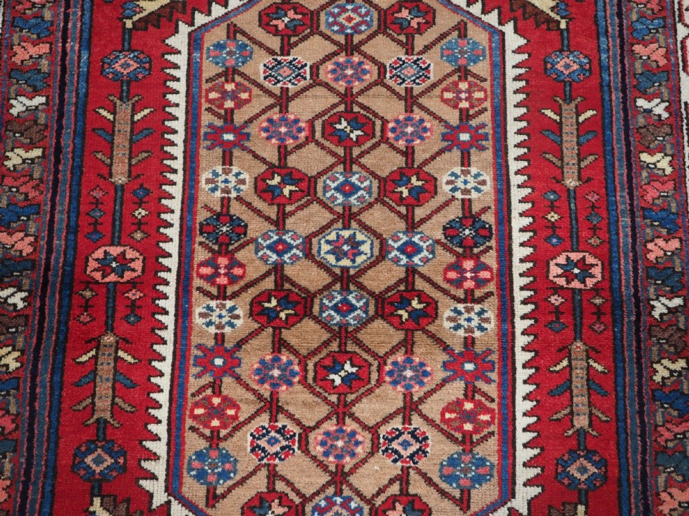 An antique hand-knotted Persian Mazlaghan wool rug featuring a central camel-coloured medallion on a red stepped field with dark blue rosette borders. View X