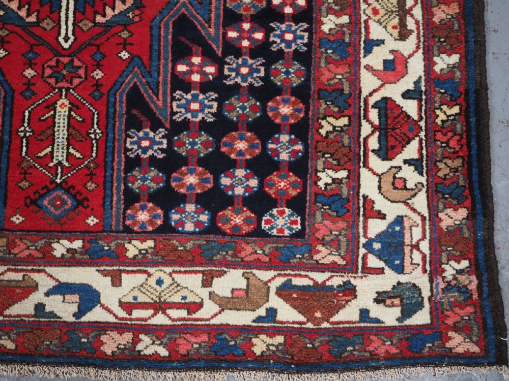 An antique hand-knotted Persian Mazlaghan wool rug featuring a central camel-coloured medallion on a red stepped field with dark blue rosette borders. View XI