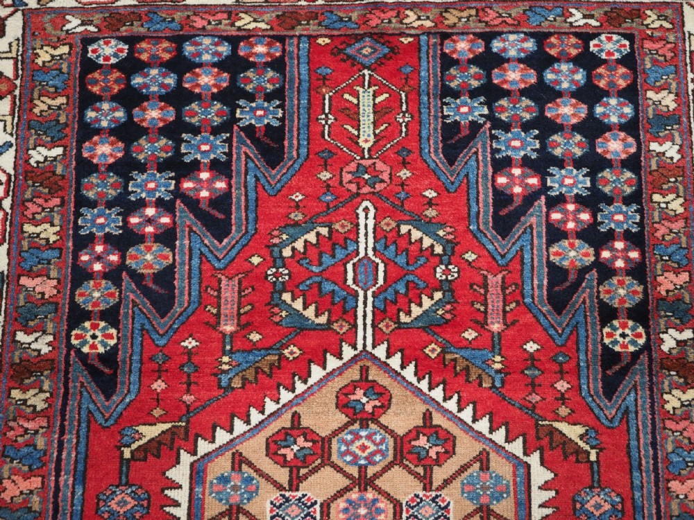 An antique hand-knotted Persian Mazlaghan wool rug featuring a central camel-coloured medallion on a red stepped field with dark blue rosette borders. View XII
