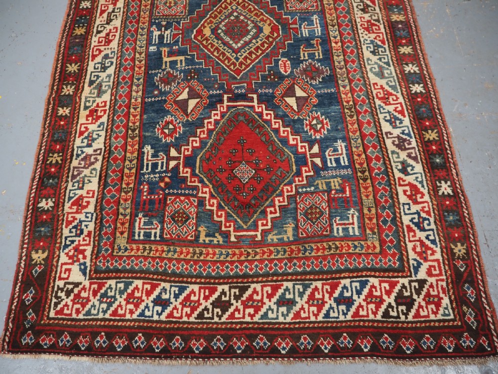 An antique Caucasian Bordjalou Kazak rug featuring a central navy blue field with two large red diamond medallions, zoomorphic animal motifs, and multi-layered geometric borders. View V