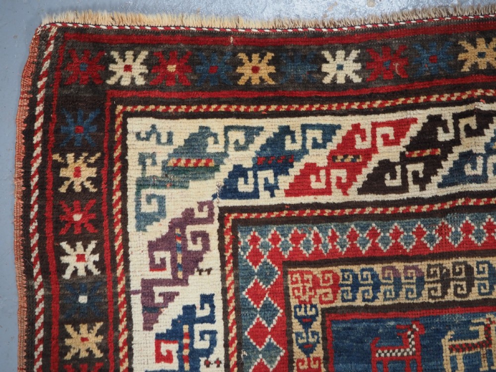An antique Caucasian Bordjalou Kazak rug featuring a central navy blue field with two large red diamond medallions, zoomorphic animal motifs, and multi-layered geometric borders. View VI