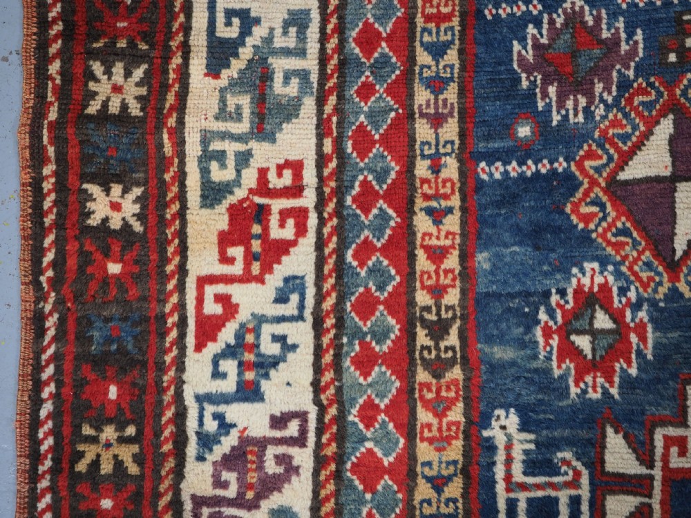 An antique Caucasian Bordjalou Kazak rug featuring a central navy blue field with two large red diamond medallions, zoomorphic animal motifs, and multi-layered geometric borders. View VII