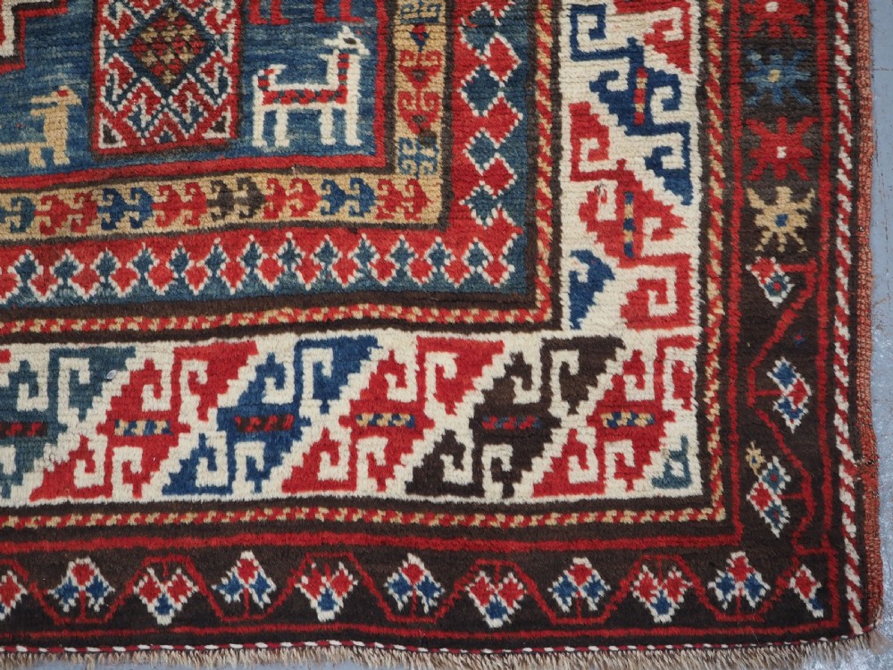 An antique Caucasian Bordjalou Kazak rug featuring a central navy blue field with two large red diamond medallions, zoomorphic animal motifs, and multi-layered geometric borders. View XV