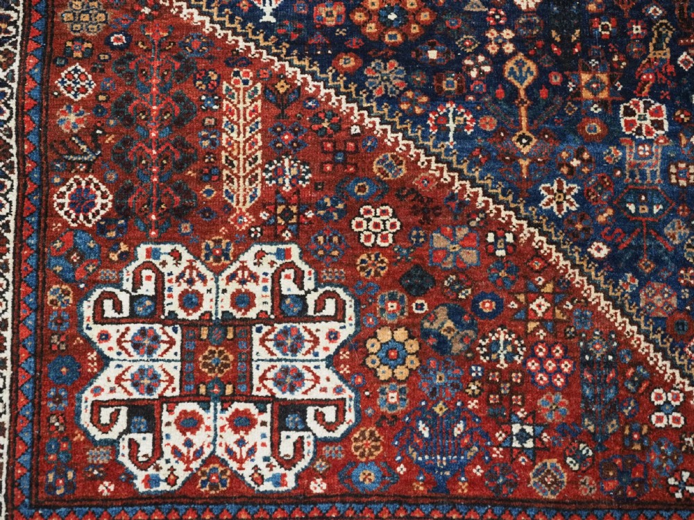 A rectangular hand-knotted antique Abadeh rug featuring a navy blue central field with a diamond medallion and rust red geometric spandrels. View X