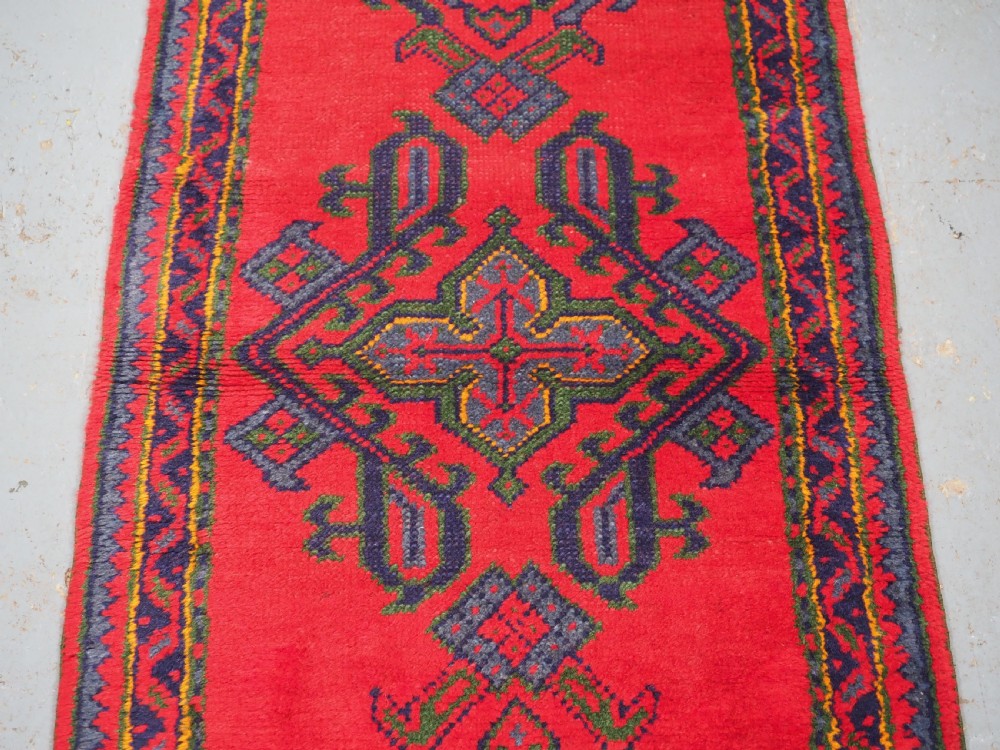 A hand-knotted red wool rug runner featuring geometric central medallions in blue, green, and yellow with a patterned border. View IV