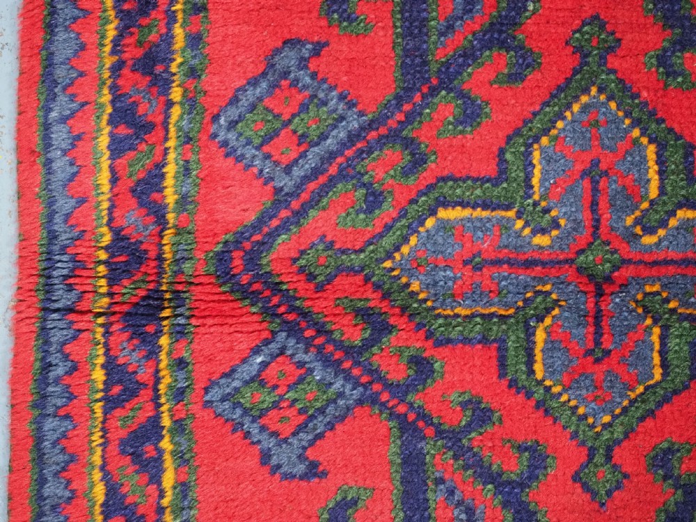 A hand-knotted red wool rug runner featuring geometric central medallions in blue, green, and yellow with a patterned border. View VII