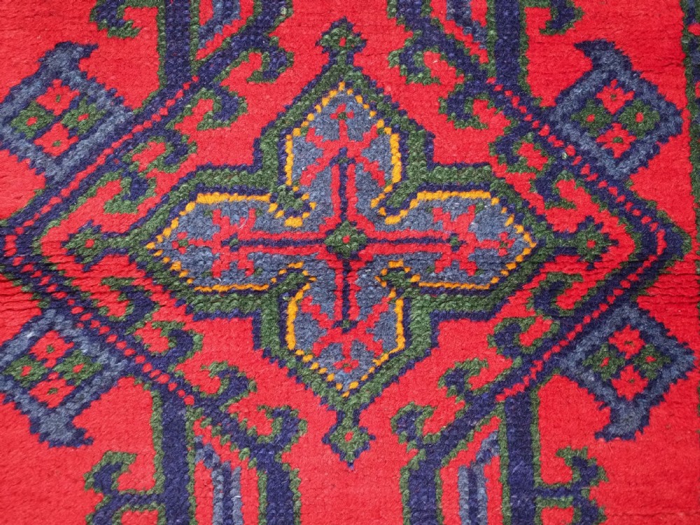 A hand-knotted red wool rug runner featuring geometric central medallions in blue, green, and yellow with a patterned border. View IX