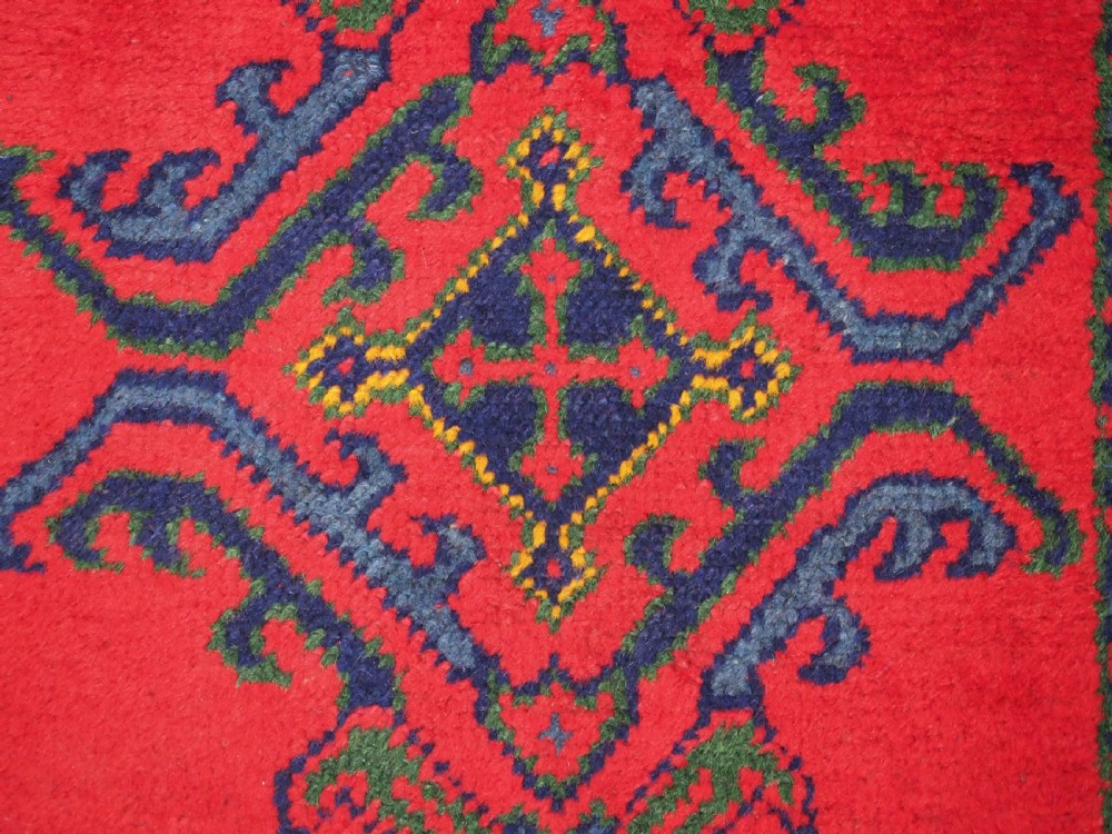 A hand-knotted red wool rug runner featuring geometric central medallions in blue, green, and yellow with a patterned border. View X