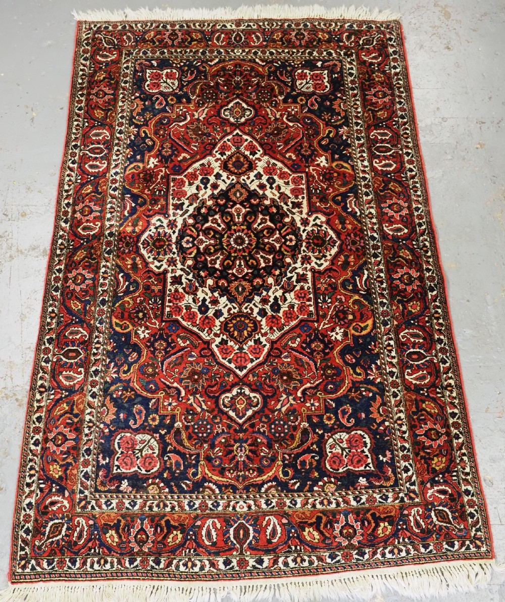 A rectangular hand-knotted Bakhtiari rug featuring a central cream floral medallion on an indigo blue field, surrounded by deep red borders and fringed edges.