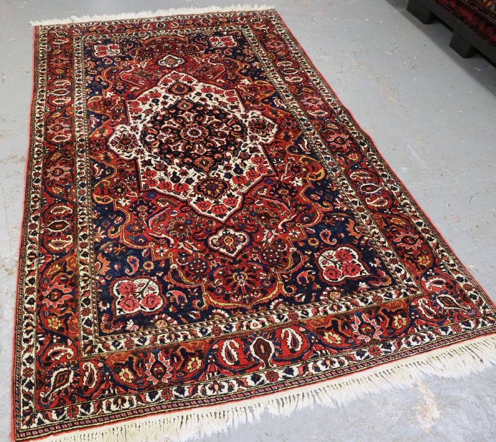 A rectangular hand-knotted Bakhtiari rug featuring a central cream floral medallion on an indigo blue field, surrounded by deep red borders and fringed edges. View II