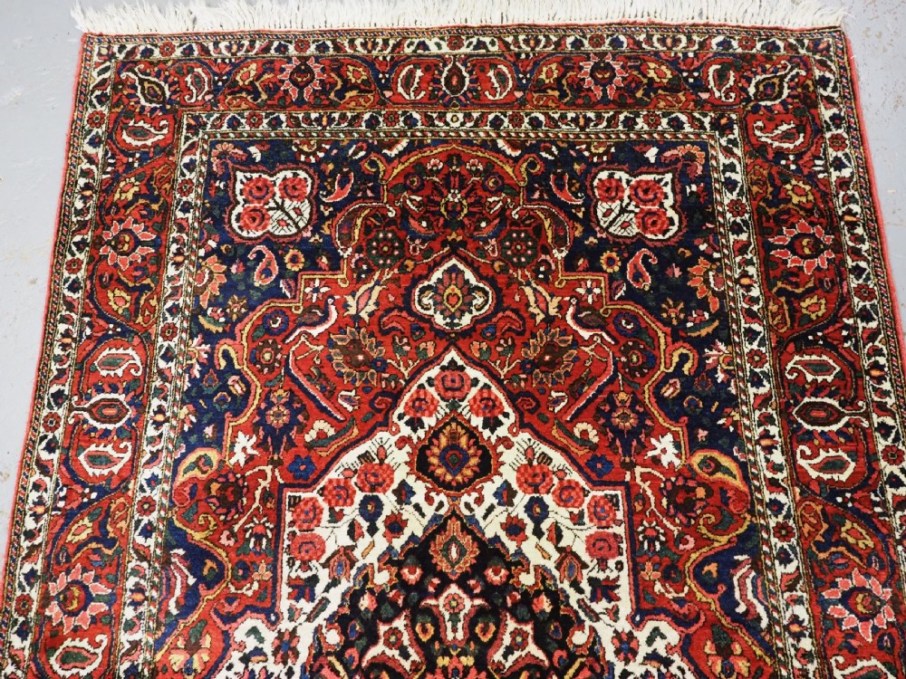 A rectangular hand-knotted Bakhtiari rug featuring a central cream floral medallion on an indigo blue field, surrounded by deep red borders and fringed edges. View III