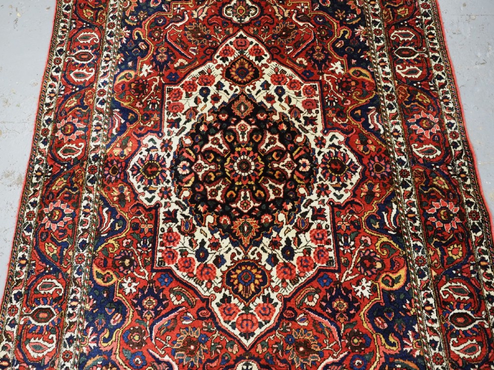 A rectangular hand-knotted Bakhtiari rug featuring a central cream floral medallion on an indigo blue field, surrounded by deep red borders and fringed edges. View IV