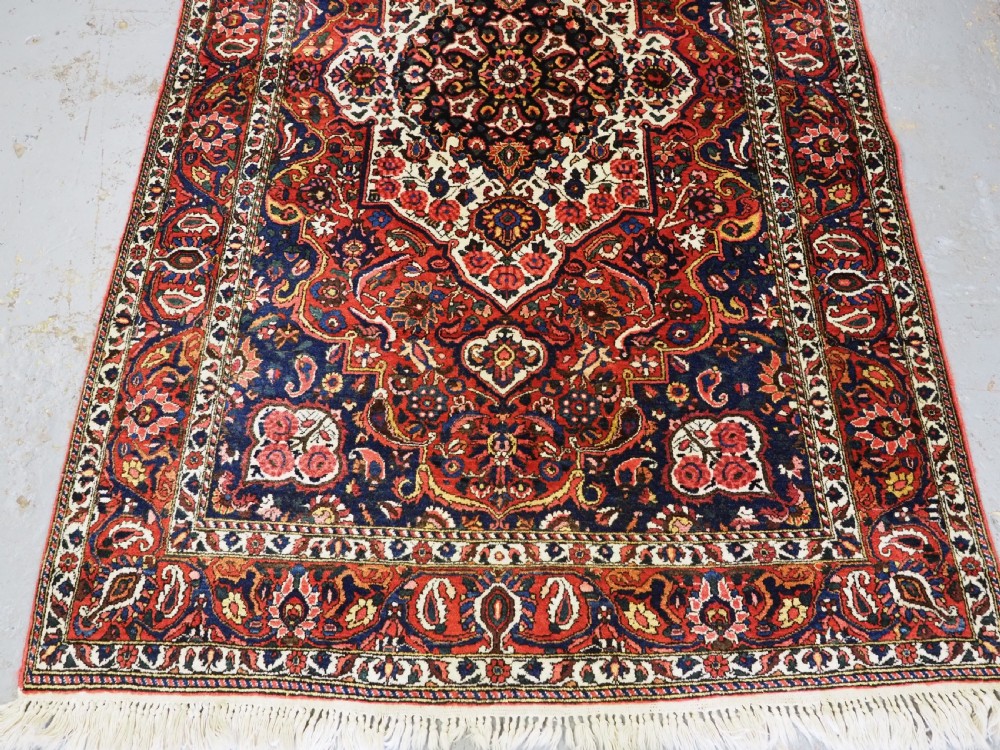 A rectangular hand-knotted Bakhtiari rug featuring a central cream floral medallion on an indigo blue field, surrounded by deep red borders and fringed edges. View V