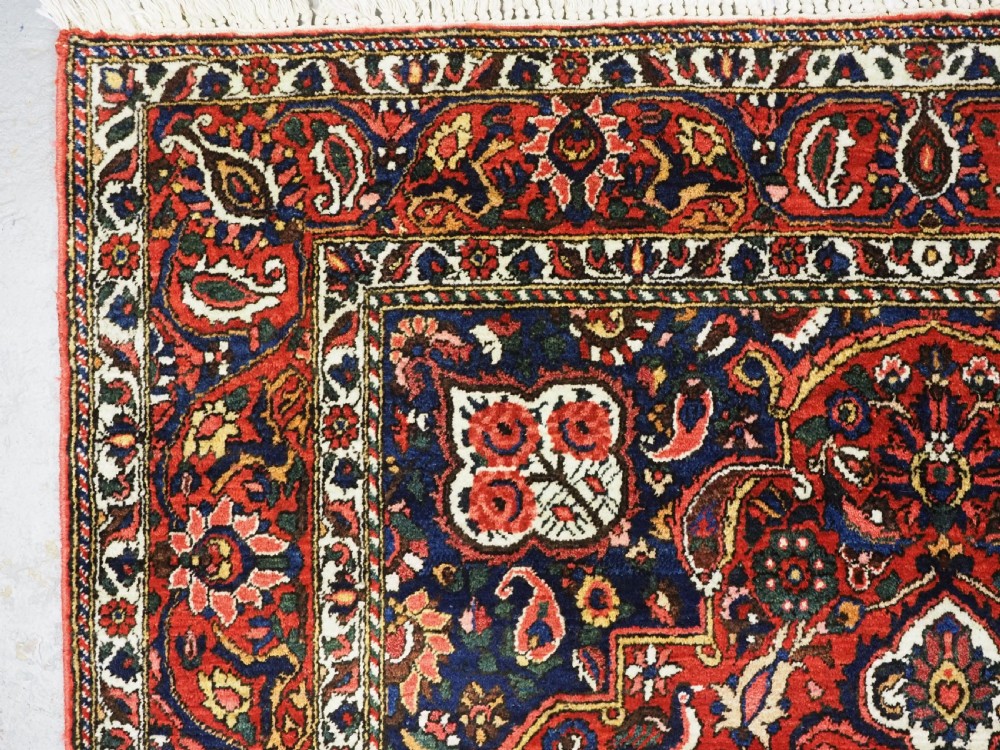 A rectangular hand-knotted Bakhtiari rug featuring a central cream floral medallion on an indigo blue field, surrounded by deep red borders and fringed edges. View VI