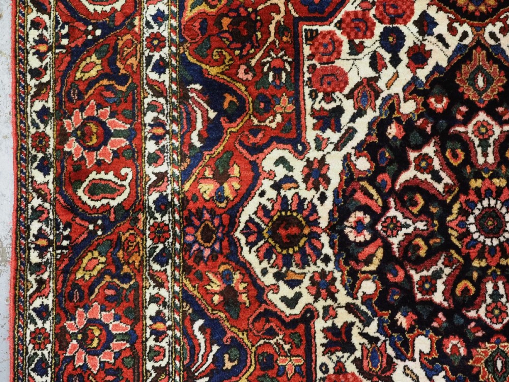 A rectangular hand-knotted Bakhtiari rug featuring a central cream floral medallion on an indigo blue field, surrounded by deep red borders and fringed edges. View VII