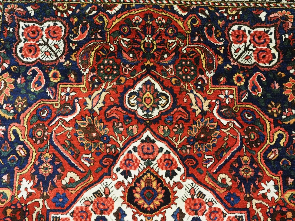 A rectangular hand-knotted Bakhtiari rug featuring a central cream floral medallion on an indigo blue field, surrounded by deep red borders and fringed edges. View VIII