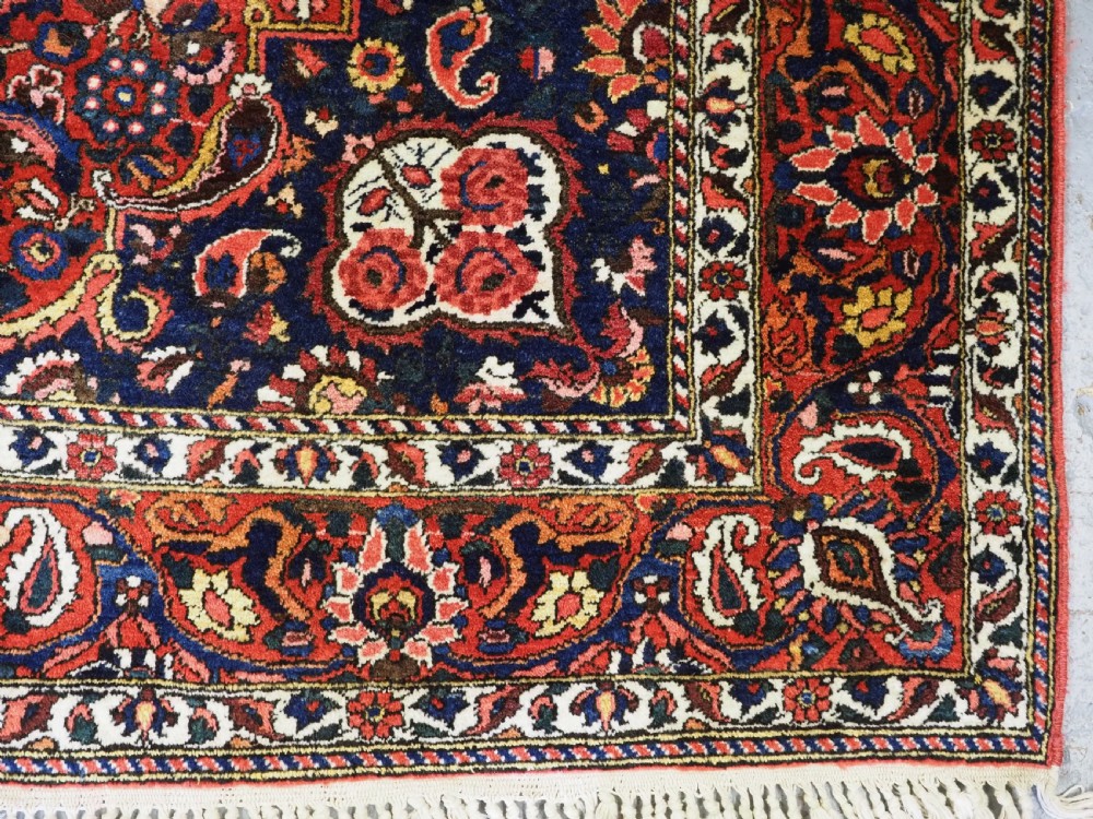 A rectangular hand-knotted Bakhtiari rug featuring a central cream floral medallion on an indigo blue field, surrounded by deep red borders and fringed edges. View IX