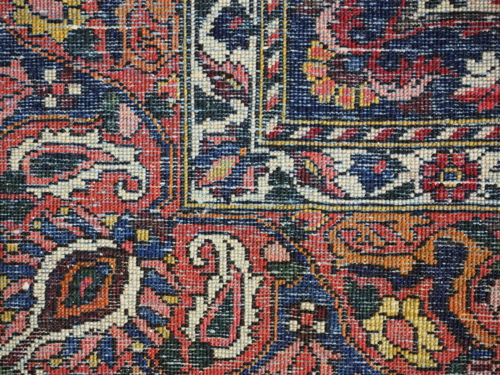 A rectangular hand-knotted Bakhtiari rug featuring a central cream floral medallion on an indigo blue field, surrounded by deep red borders and fringed edges. View X