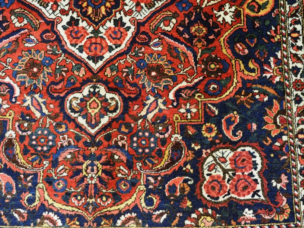 A rectangular hand-knotted Bakhtiari rug featuring a central cream floral medallion on an indigo blue field, surrounded by deep red borders and fringed edges. View XI