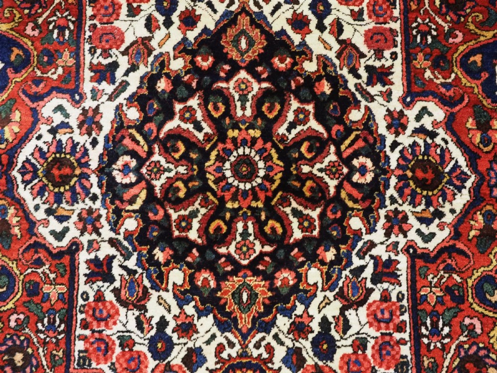 A rectangular hand-knotted Bakhtiari rug featuring a central cream floral medallion on an indigo blue field, surrounded by deep red borders and fringed edges. View XII