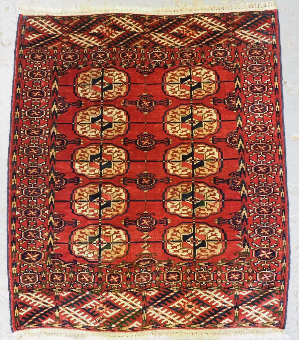 A small rectangular Tekke Turkmen hand-knotted wool rug featuring two vertical columns of geometric octagonal guls on a deep red ground with patterned borders and fringed edges.