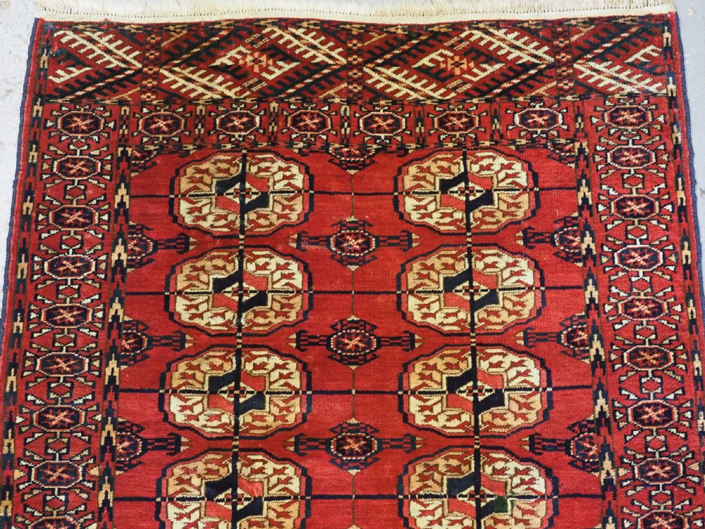 A small rectangular Tekke Turkmen hand-knotted wool rug featuring two vertical columns of geometric octagonal guls on a deep red ground with patterned borders and fringed edges. View III