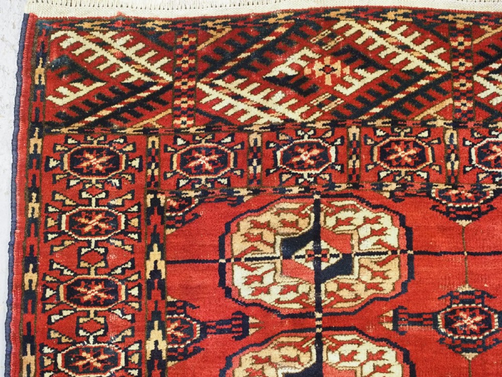 A small rectangular Tekke Turkmen hand-knotted wool rug featuring two vertical columns of geometric octagonal guls on a deep red ground with patterned borders and fringed edges. View IV
