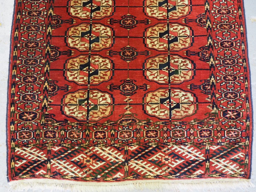 A small rectangular Tekke Turkmen hand-knotted wool rug featuring two vertical columns of geometric octagonal guls on a deep red ground with patterned borders and fringed edges. View V