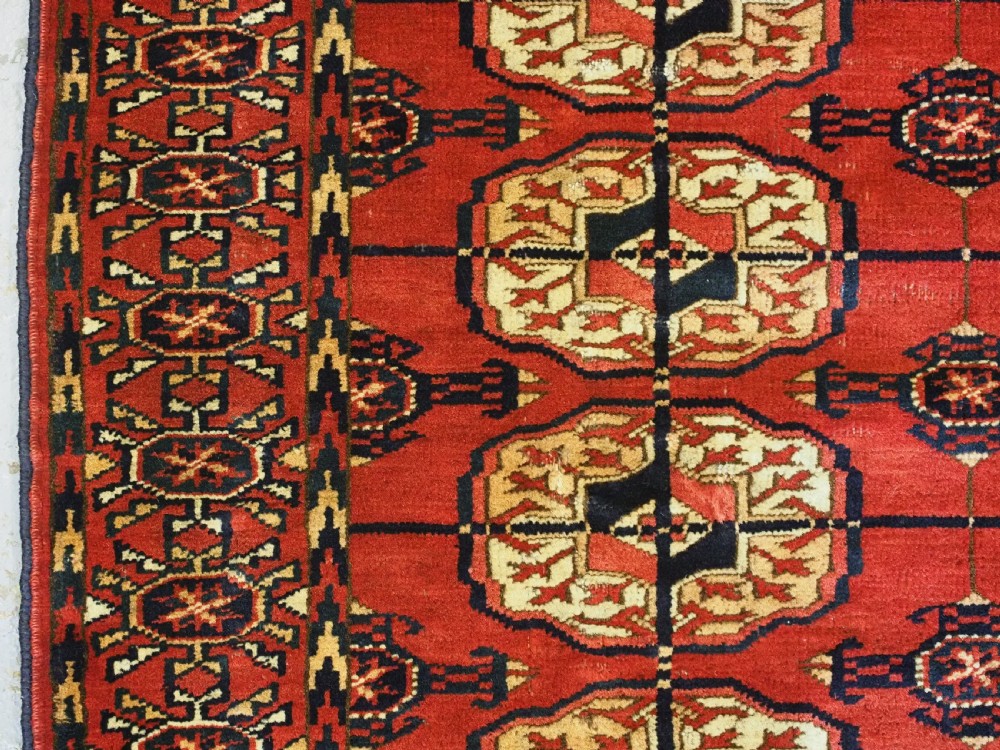 A small rectangular Tekke Turkmen hand-knotted wool rug featuring two vertical columns of geometric octagonal guls on a deep red ground with patterned borders and fringed edges. View VI