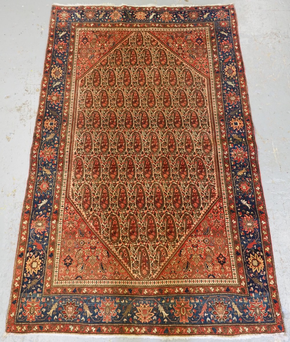 An antique Persian Sarouk rug featuring a repeating central boteh pattern framed by detailed floral borders in shades of terracotta red, cream, and navy blue.