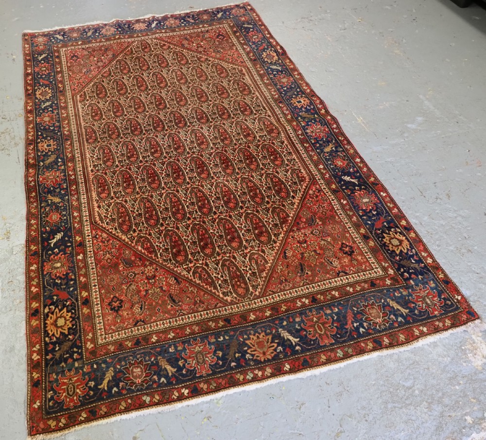 An antique Persian Sarouk rug featuring a repeating central boteh pattern framed by detailed floral borders in shades of terracotta red, cream, and navy blue. View III
