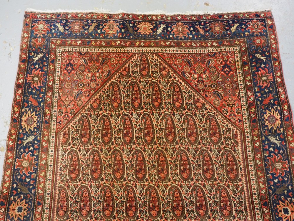 An antique Persian Sarouk rug featuring a repeating central boteh pattern framed by detailed floral borders in shades of terracotta red, cream, and navy blue. View IV