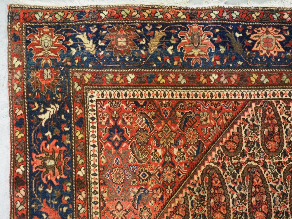 An antique Persian Sarouk rug featuring a repeating central boteh pattern framed by detailed floral borders in shades of terracotta red, cream, and navy blue. View VII