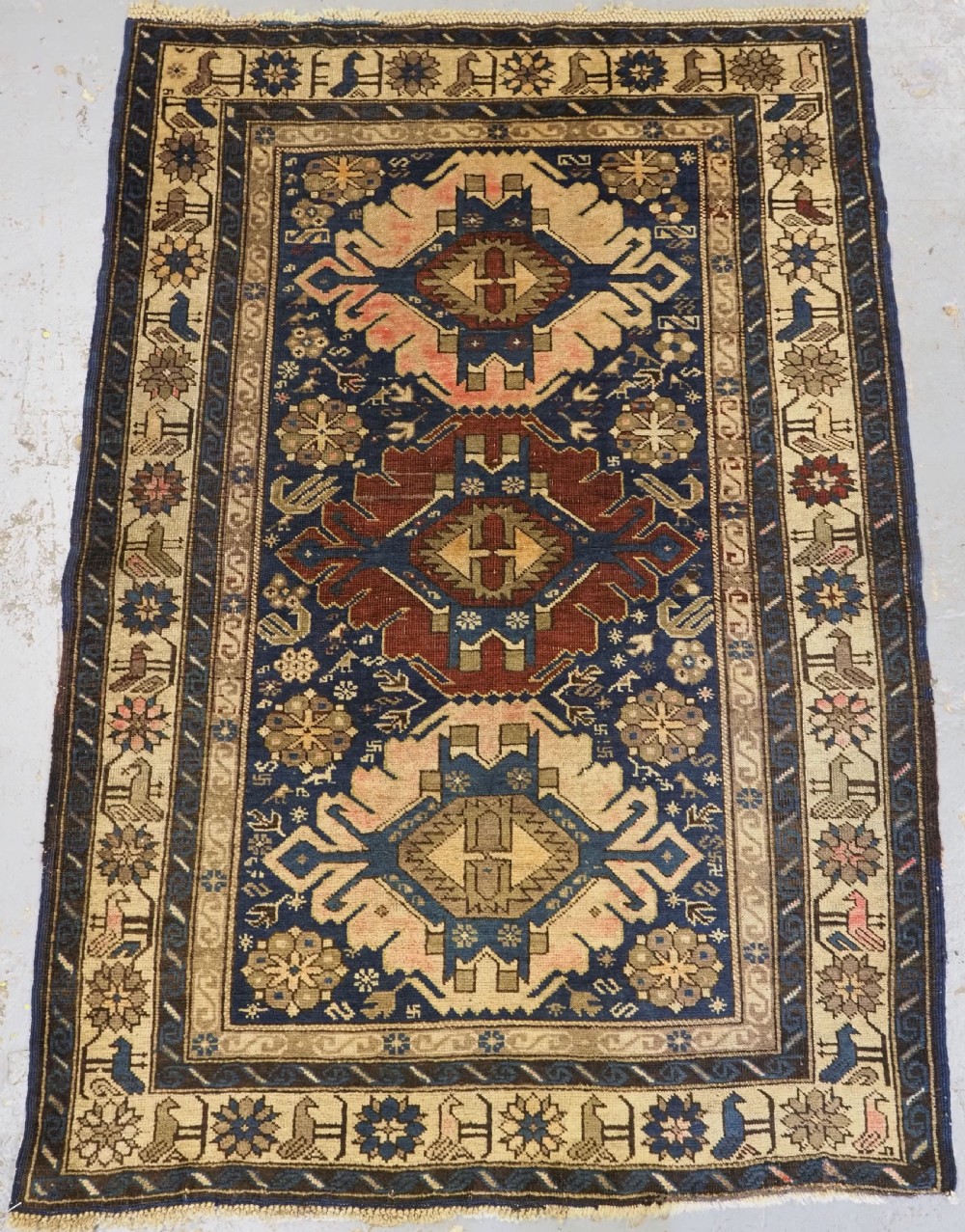 An antique hand-knotted Caucasian rug featuring three central geometric medallions on a navy blue field with a beige border adorned with stylized bird motifs.