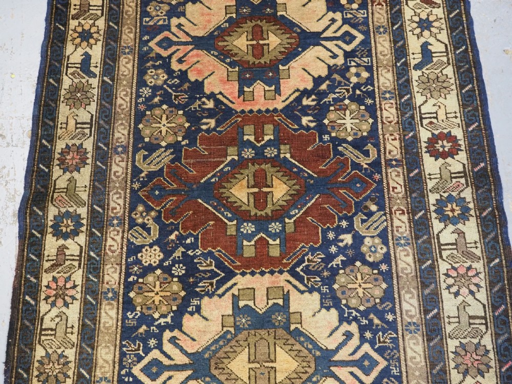 An antique hand-knotted Caucasian rug featuring three central geometric medallions on a navy blue field with a beige border adorned with stylized bird motifs. View IV