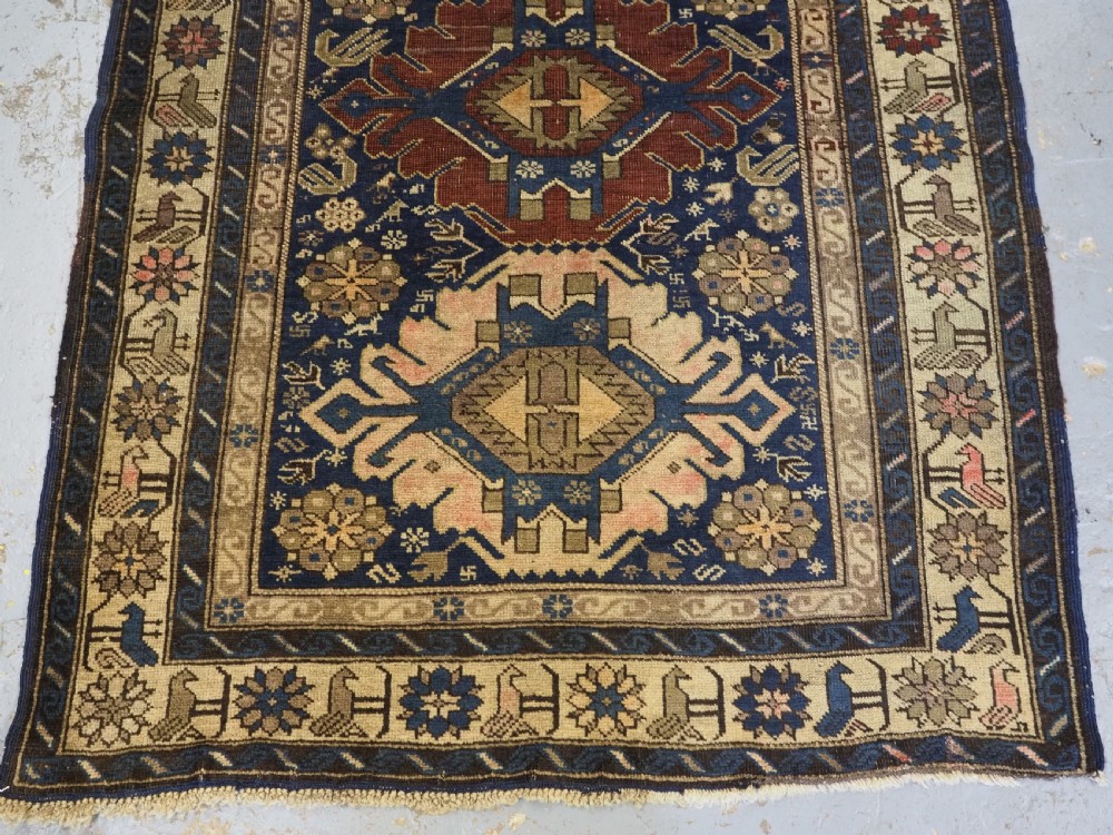 An antique hand-knotted Caucasian rug featuring three central geometric medallions on a navy blue field with a beige border adorned with stylized bird motifs. View V