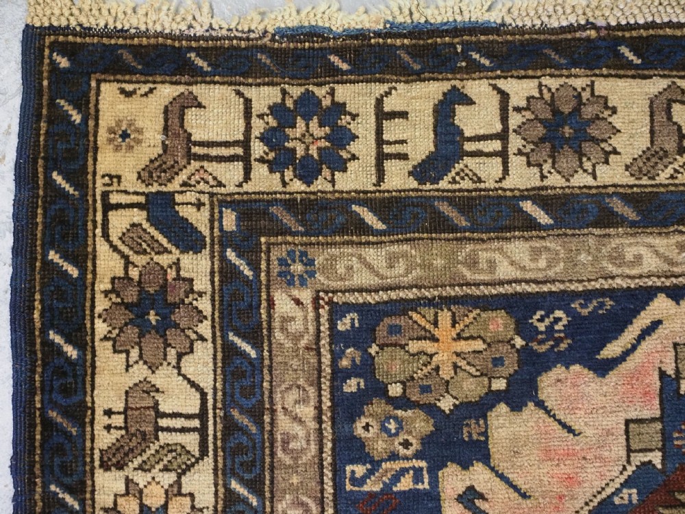 An antique hand-knotted Caucasian rug featuring three central geometric medallions on a navy blue field with a beige border adorned with stylized bird motifs. View VI