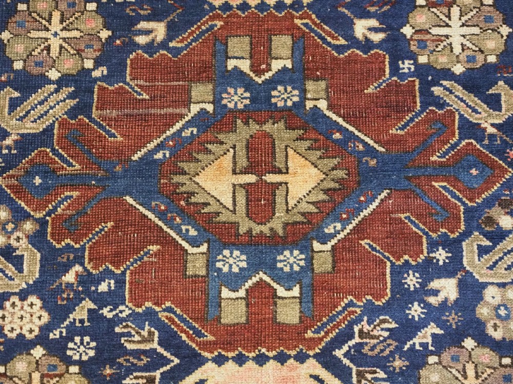 An antique hand-knotted Caucasian rug featuring three central geometric medallions on a navy blue field with a beige border adorned with stylized bird motifs. View IX