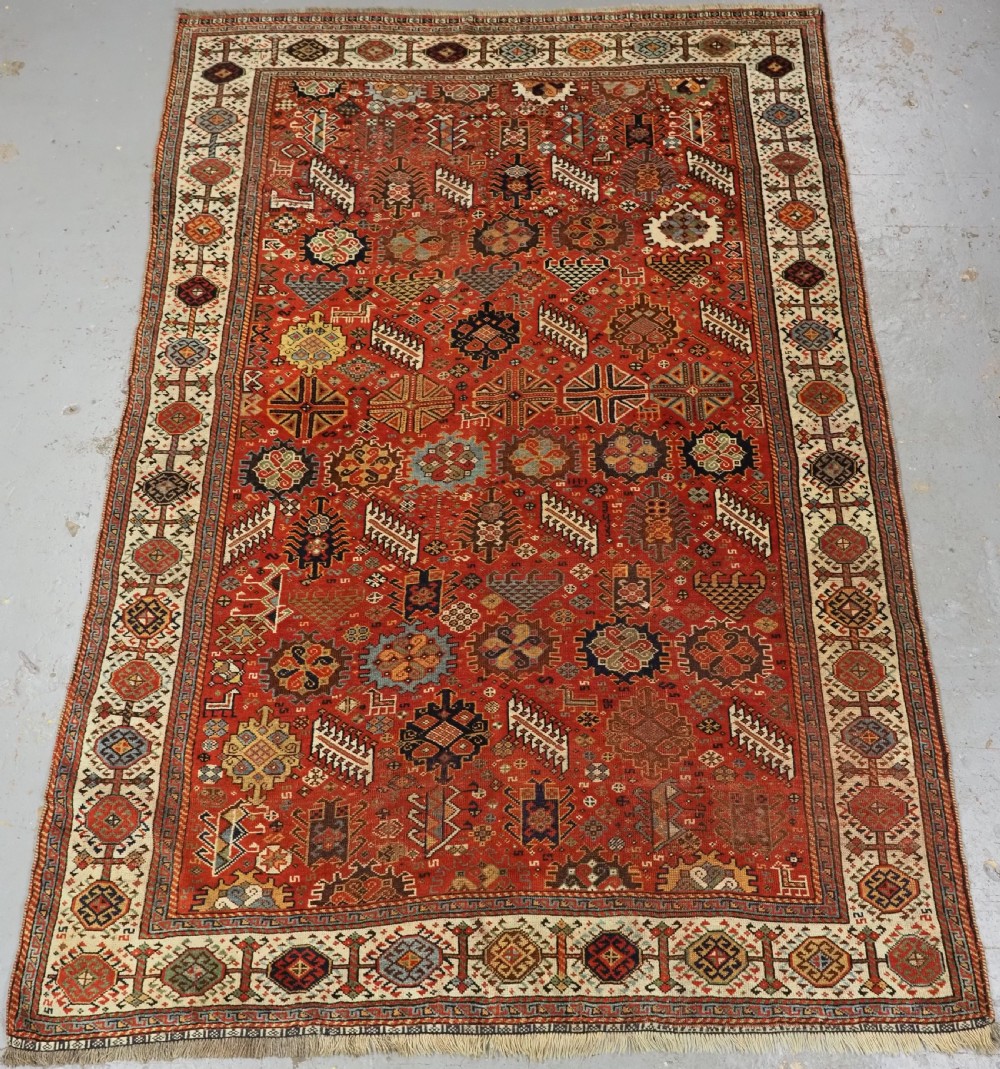 An antique South Persian Shekarlu Qashqai wool rug featuring a red field with dense geometric tribal motifs surrounded by an ivory patterned border.