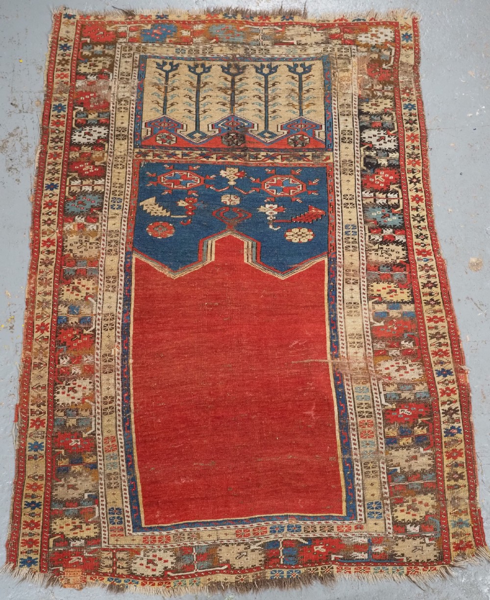 An antique hand-knotted Turkish Ladik prayer rug featuring a central red arched mihrab niche, blue spandrels, stylized floral panels, and a patterned multi-tiered border.