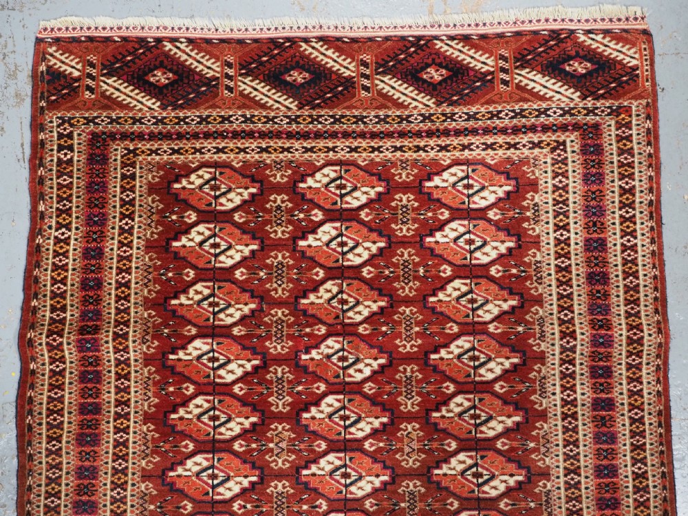 A rectangular handwoven Tekke Turkmen wool rug featuring repeating primary gul motifs in deep red, navy, terracotta, and ivory tones surrounded by geometric borders. View III