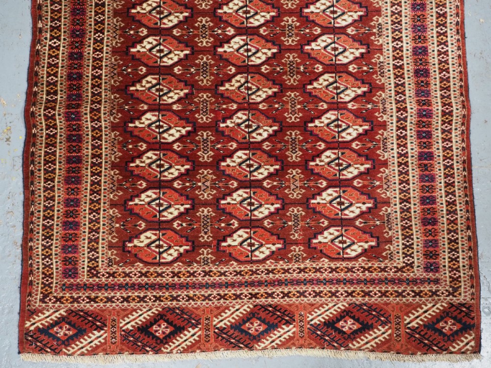 A rectangular handwoven Tekke Turkmen wool rug featuring repeating primary gul motifs in deep red, navy, terracotta, and ivory tones surrounded by geometric borders. View IV