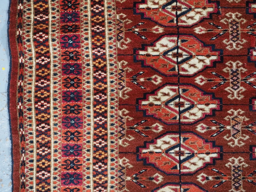 A rectangular handwoven Tekke Turkmen wool rug featuring repeating primary gul motifs in deep red, navy, terracotta, and ivory tones surrounded by geometric borders. View VI